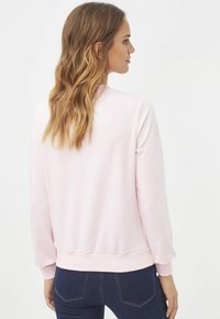 Sudadera rosa claro con mangas largas, puños y dobladillo acanalados, textura suave y un ajuste relajado. Llevada sobre jeans oscuros, vista desde la parte posterior.