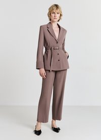 Brauner maßgeschneiderter Blazer mit einem Taillengürtel, zwei frontalen Taschen und schwarzen Knöpfen, kombiniert mit passenden geraden Hosen und schwarzen Schuhen.