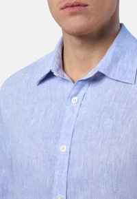 Camicia in lino azzurro chiaro con colletto all'inglese, bottoni bianchi, tessuto testurizzato e un piccolo logo sul petto. Vista ravvicinata del collo.