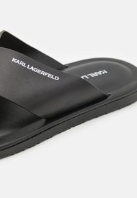 Melna slaidu sandalīte ar divām platajām, nedaudz strukturētām siksnām. Zīmola nosaukums "KARL LAGERFELD" ir izdrukāts baltā krāsā uz siksnas.