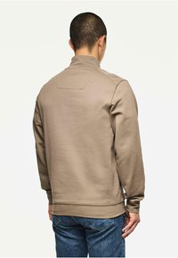 Beige sweatshirt met hoge kraag, ribgebreide manchetten en onderrand. Bevat een rechthoekig schouderdetail op de achterkant. Gedoofd met blauwe spijkerbroeken.