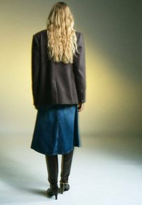 Przesadnie duża marynarka w kratę w ciemnych odcieniach, zestawiona z luźną niebieską spódnicą dżinsową i brązowymi kozakami do kolan z szpiczastymi noskami.