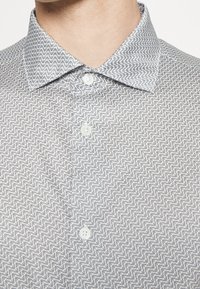 Close-up of a person wearing a light gray patterned button-up shirt with a pointed collar and three visible white buttons.