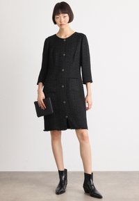 Abito in tweed nero con maniche a tre quarti, chiusura con bottoni, due tasche e orlo sfrangiato. Abbinato a stivali neri alla caviglia e una pochette.
