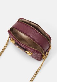 Bordeauxfarbene Stepp-Handtasche mit goldenen Beschlägen, die über zwei Reißverschlussfächer und einen Kettenriemen verfügt. Das Innenfutter ist beige mit einem dezenten Muster.