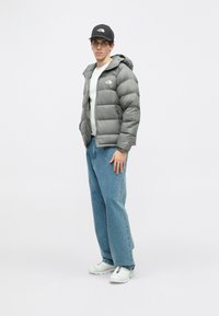 Szürke puffermellény kapucnival, fehér ing felett viselve. Laza kék farmer és fehér sportcipő egészíti ki a megjelenést, fekete sapka a fejen.