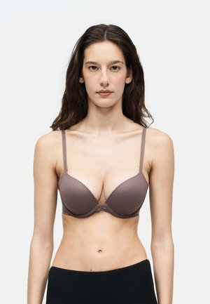 Young woman with long dark hair wearing a mauve bra and black pants, facing forward against a plain white background.