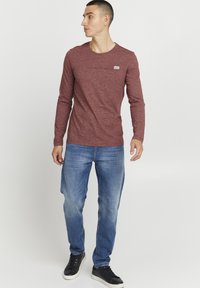 Langermet maroon t-skjorte med rund hals og brystlomme, kombinert med lyseblå jeans og svarte joggesko.