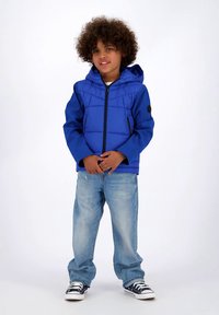 Copil tânăr cu păr creț, purtând o jachetă albastră cu glugă, blugi bleu deschis și adidași alb-negru, stând în picioare pe un fundal simplu.