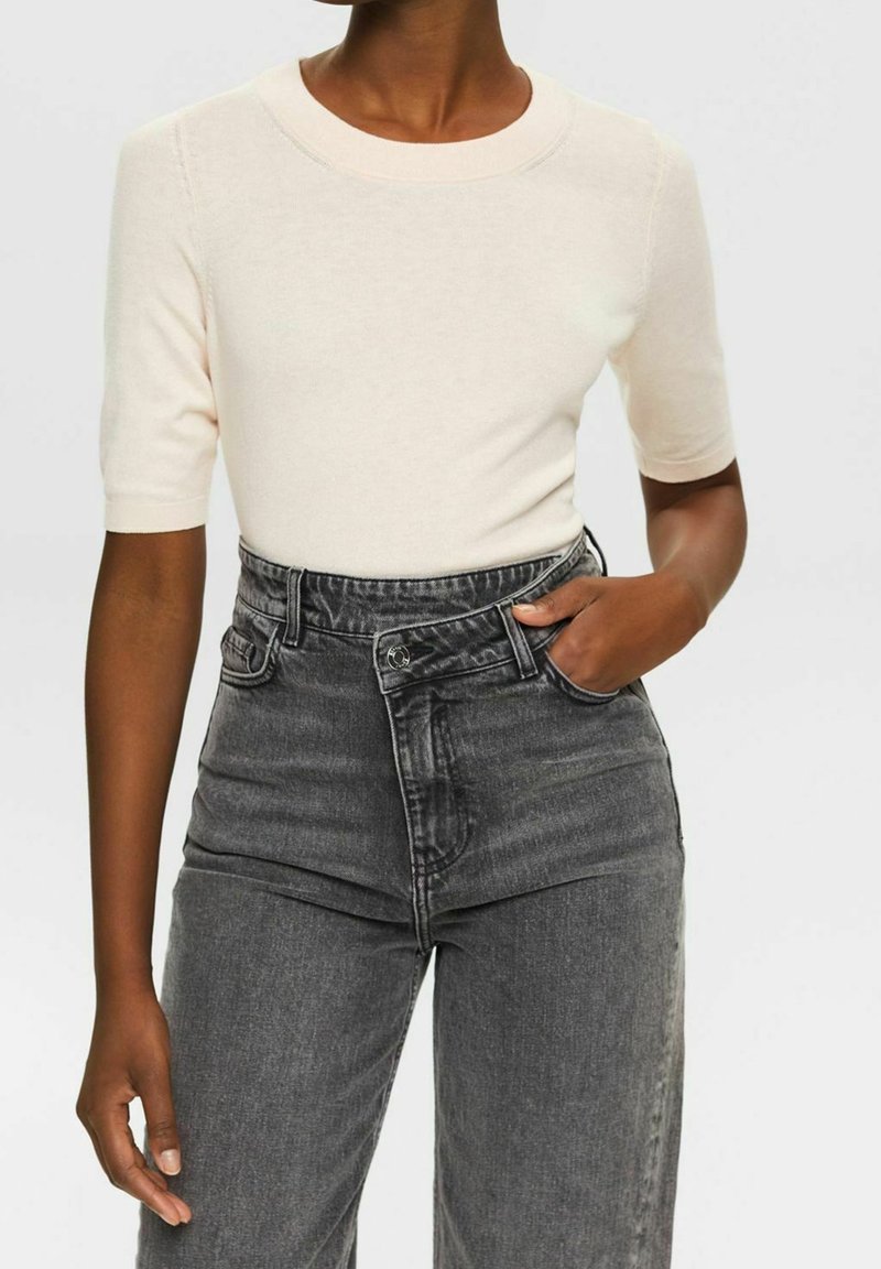 Maglione corto a maniche corte di colore crema chiaro con scollo rotondo, abbinato a jeans in denim grigio scuro a vita alta, leggermente usurati.