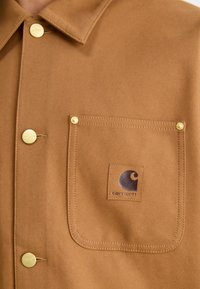 Giacca a camicia marrone realizzata in morbido tessuto, con una tasca sul petto decorata con il logo Carhartt, bottoni a pressione dorati e cuciture a contrasto.