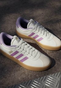 Zapatillas de cuero blancas con rayas moradas, suela de goma texturizada, collar acolchado y cordones en tono, con un logo dorado en el lateral.