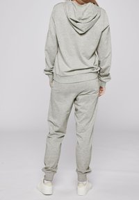 Person som står med händerna i fickorna, iförd ljusgrå huvtröja och matchande sweatpants, vita sneakers, vänd bort från kameran mot en enfärgad bakgrund.