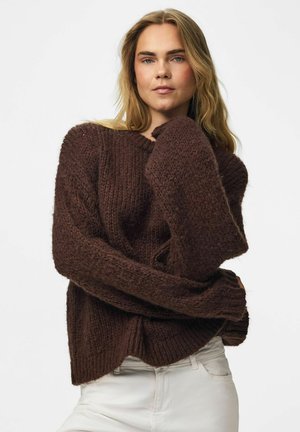 Kvinde med langt blond hår iført en chunky mørkebrun strikket sweater og lyse bukser, står med armene krydset mod en ensfarvet baggrund.