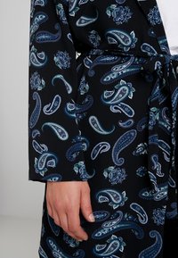 Svart klänning med ett paisley- och blommönster i blå och gröna toner, utrustad med bälte och långa ärmar. Tyget verkar slätt och lätt.