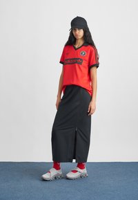 Moteris vilkinti raudoną "Adidas" sportinę marškinėlį, juodą kepurę, ilgą juodą sijoną su šoniniu užtrauktuku, raudonas kojines ir pilkas sportines batus, stovinti ant mėlyno grindų.