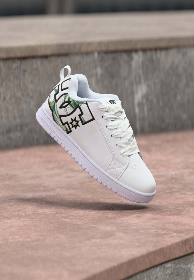 Λευκό sneaker χαμηλού ύψους με λευκά κορδόνια και μαύρο-πράσινο σχέδιο γραφικών στην πλευρά, να αιωρείται πάνω από μια ροζ πέτρινη επιφάνεια.