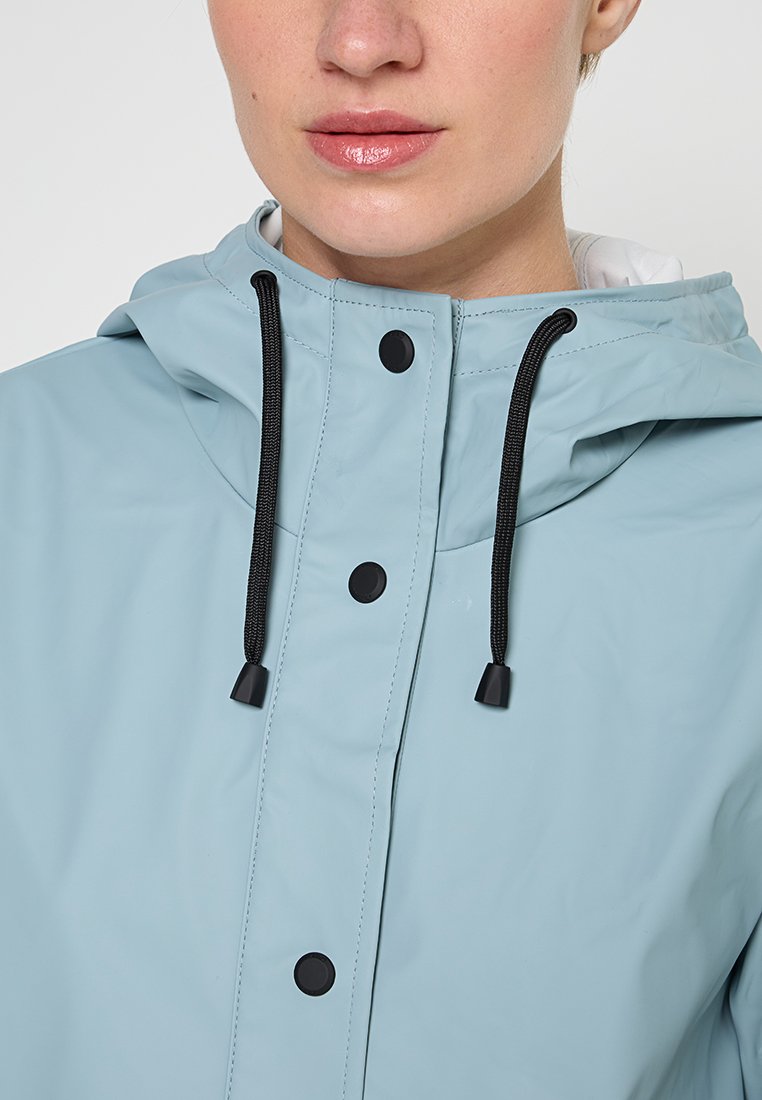 Chaqueta impermeable azul claro con capucha, con cordones negros, botones y una textura suave. Primer plano de los detalles frontales.