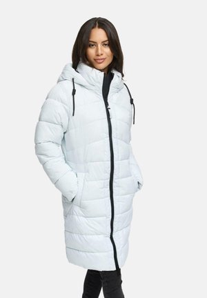 Woman wearing a long light blue padded winter coat with hood and black zipper, hands in pockets, standing against a plain background.