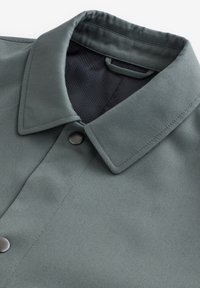 Chaqueta verde oscuro con cuello, botones de presión y forro interior de malla negra, mostrada en detalle de cerca.