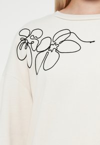 Lichtbeige sweatshirt met zwarte bloemen borduursel op de linkerschouder. Zachte textuur en een ontspannen pasvorm met een ronde hals.