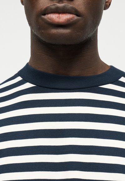 Κοντινό πλάνο στο κάτω μέρος του προσώπου και τον λαιμό ενός ατόμου που φοράει μπλούζα με οριζόντιες ρίγες σε σκούρο μπλε και λευκό χρώμα, με στρογγυλή λαιμόκοψη.