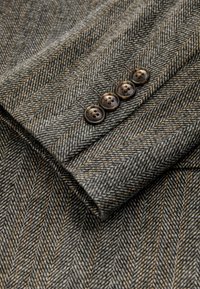 Gray herringbone wool fabric jacket cuff featuring four circular brown buttons with floral designs, showing a textured, tailored finish.