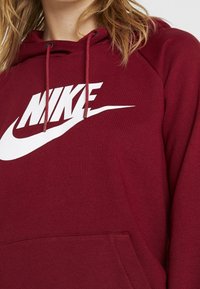 Kvinna i en mörkröd huvtröja med en vit Nike-logotyp och snörning, delvis visad från halsen till midjan.