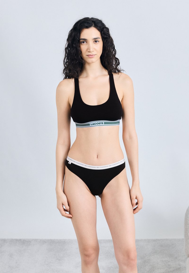 Woman with curly hair wearing black Lacoste sports bra and matching underwear standing against a plain light gray background.