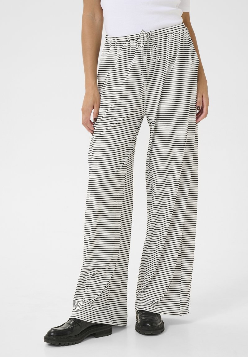 Personne portant un pantalon ample à rayures horizontales noires et blanches avec une taille à cordon et des chaussures noires épaisses, debout sur un sol blanc.