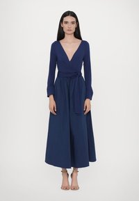 Marineblauwe langemouwen wikkeldress, diepe V-hals, taille cinch met een strik, vloeiende rok, zachte stof en enkellange schoenen in een neutrale tint.