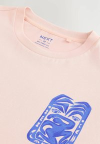 Camiseta de algodón rosa claro con un corte relajado; presenta un diseño gráfico azul que representa formas abstractas y rasgos faciales en la parte frontal.