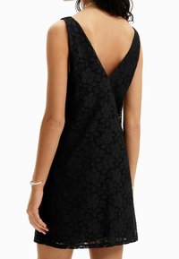 Robe en dentelle noire avec un dos en V, sans manches, motif floral et ourlet légèrement évasé. Elle présente une texture douce et une silhouette féminine.