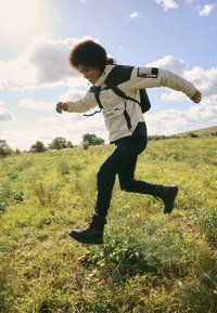 Beige wasserdichte Jacke mit schwarzen Akzenten, schwarze Hose und robuste schwarze Stiefel. Das Motiv springt in einem grasbewachsenen Feld unter einem blauen Himmel.