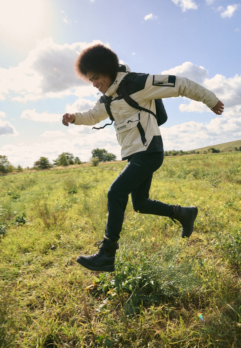 Chaqueta impermeable beige con detalles en negro, pantalones negros y botas negras resistentes. El sujeto está saltando en un campo de hierba bajo un cielo azul.
