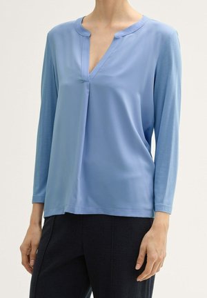 Kobieta ubrana w jasnoniebieską bluzkę z długim rękawem i dekoltem w serek z plisowanym detalem, w połączeniu z czarnymi spodniami o fakturze.