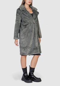 Cappotto grigio in pelliccia sintetica con design doppiopetto, colletto a pistagna e tasche laterali. Il modello indossa stivali neri con zip alla caviglia.