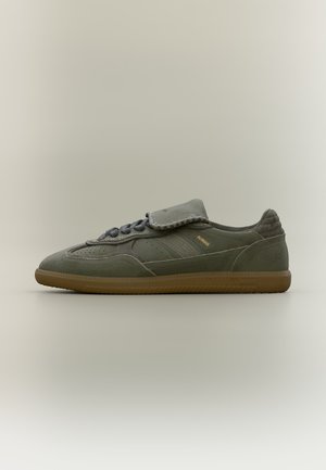 Alacsony profilú olíva zöld nubuk sneaker gumitalppal, elöl fűzős, meghosszabbított nyelvredővel, és finom arany logóval az oldalon.