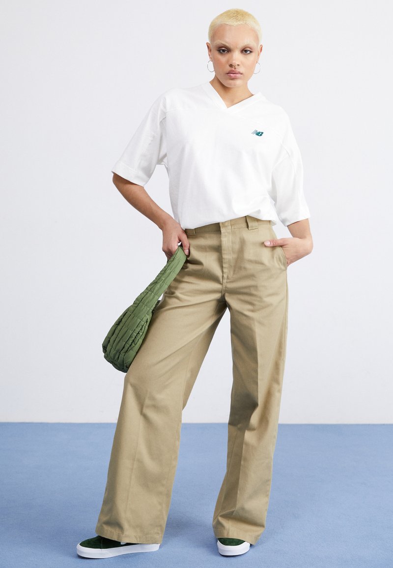 Weißes, übergroßes V-Ausschnitt-T-Shirt, beige, weite Hose, grüne, strukturierte Tasche und grüne Sneakers, vor einem hellblauen Boden und weißen Hintergrund.