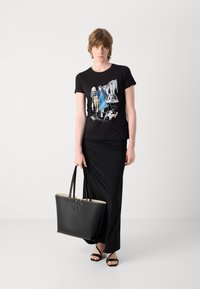 Black graphic t-shirt featuring a printed design, paired with a black satin skirt. Holding a large black tote bag with embossed "DKNY" logo, and black sandals.