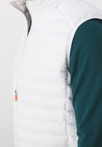 Hvid quiltet vest med høj krave og lynlåsfortil. Har en lille orange detalje på lynlåsen og bæres over en teal-farvet skjorte.
