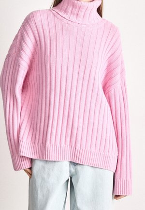 Personne portant un pull à col roulé côtelé rose oversize associé à un jean bleu clair sur un fond uni.