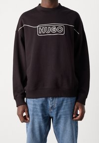 Sudadera negra con cuello y puños acanalados, que presenta un bordado blanco del logo "HUGO" en la parte frontal. Ajuste regular con mangas largas.