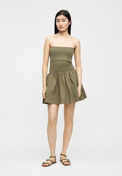 Νεαρή γυναίκα με μακριά σκουρόχρωμα μαλλιά, φορώντας αμάνικο φόρεμα σε ελιά πράσινο και καφέ επίπεδα σανδάλια, στέκεται μπροστά σε απλό φωτεινό φόντο.