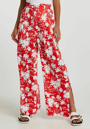 Rode broek met wijde pijpen en een wit bloemenpatroon, elastische taille met trekkoord, zijsplitten, gedragen met witte sandalen en een witte top.