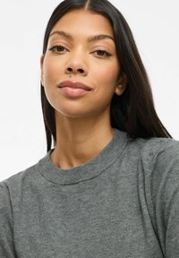 Sweatshirt gris avec un col rond côtelé. Le modèle a de longs cheveux foncés et une peau lisse. Pas de motifs ni de logos visibles. Design simpliste.