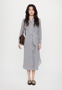 Abito a righe con bottoni blu e bianco, con vita cintata, maniche lunghe e orlo asimmetrico. La modella tiene in mano una borsa a tracolla marrone.