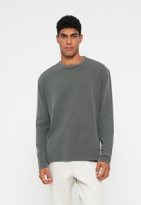 Mann, der vor einem weißen Hintergrund steht, trägt ein graues Langarmshirt und hellfarbene Hosen, blickt nach vorne mit einem neutralen Ausdruck.