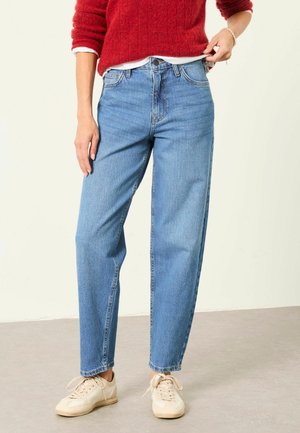 Person iført lyseblå jeans med lige ben, rød sweater, hvide sneakers og justerer sømmen på sweateren med den ene hånd.