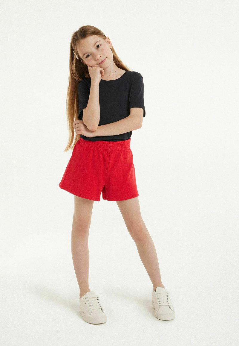 Haut noir à manches courtes, short rouge avec une taille élastique et baskets blanches. Design simple, texture lisse et couleurs vives.